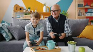 Grandfather and grandson are playing video game sitting on sofa at home using joysticks enjoying entertainment. Family and modern technology concept.