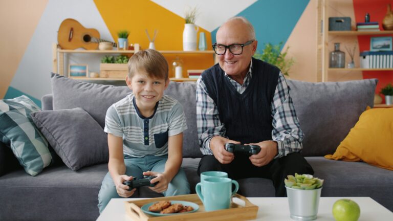 Grandfather and grandson are playing video game sitting on sofa at home using joysticks enjoying entertainment. Family and modern technology concept.